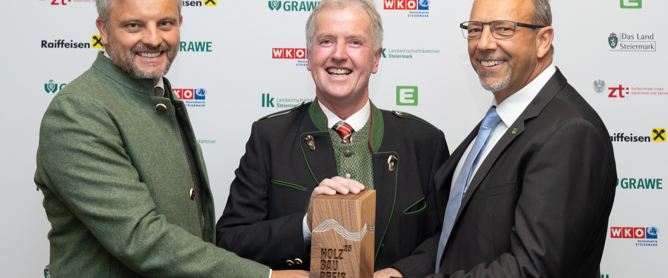 Drei Männer in Anzügen und Tracht lächeln in die Kamera und halten gemeinsam eine Holz-Trophäe mit der Aufschrift 