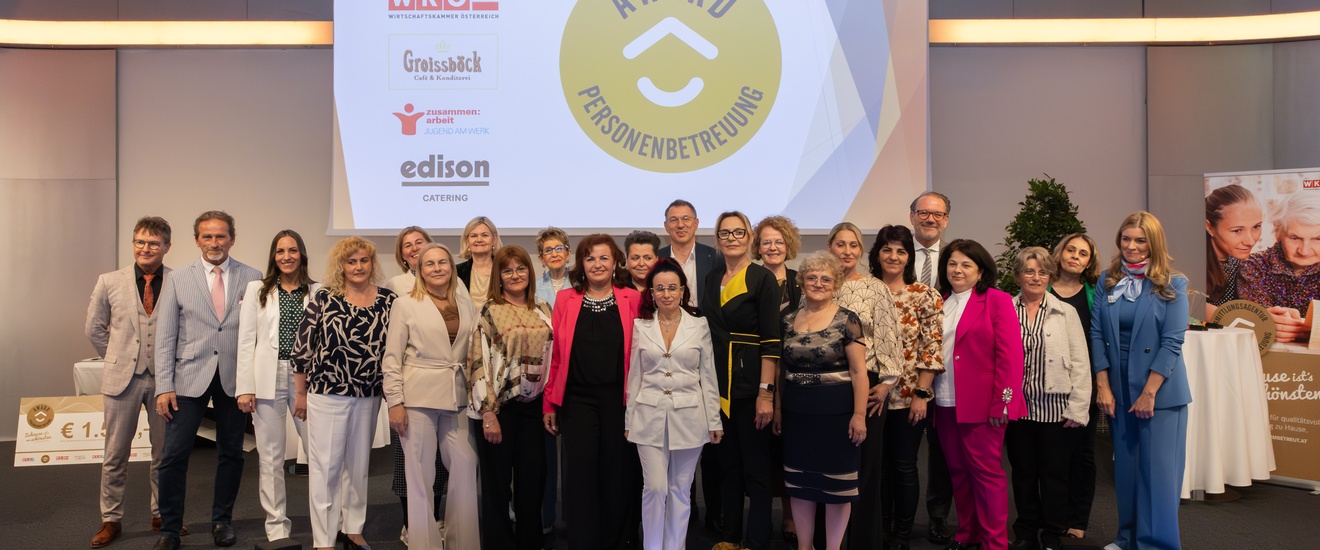 Eine Gruppe von rund 20 Personen steht nebeneinander auf einer Bühne und blickt in die Kamera. Im Hintergrund ist eine große Projektionsfläche mit einem runden gelben Logo und dem Text „AWARD PERSONENBETREUUNG“