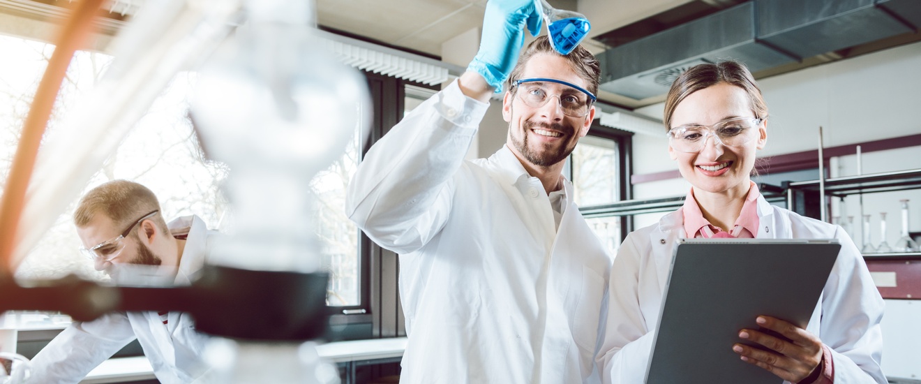 Drei Personen in weißen Kitteln mit Schutzbrillen in Labor, eine Person schwenkt Glas mit Flüssigkeit, eine andere blickt auf Clipboard