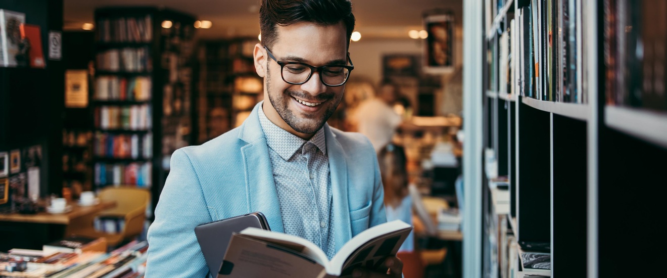 Lächelnde Person mit Brillen steht in Buchhandlung und blickt in aufgeschlagenes Buch