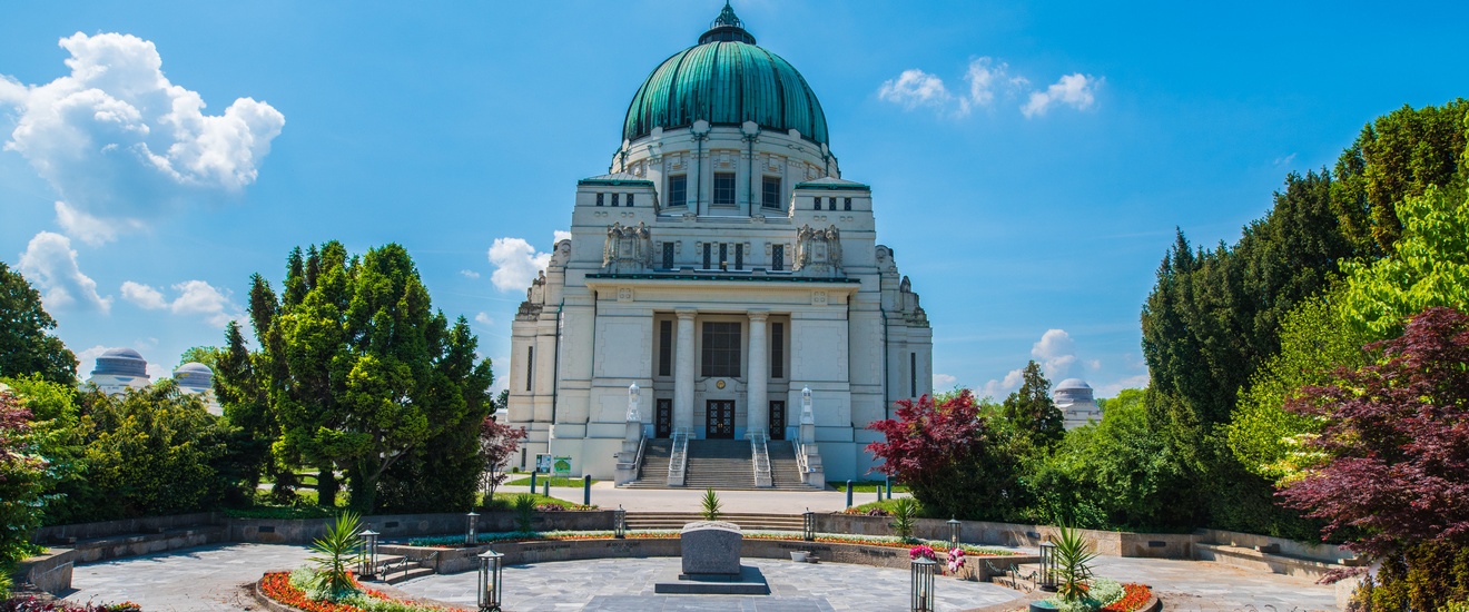 In der Mitte ist ein runder Platz mit einem steinern Sarg. Links und rechts davon sind grüne Büsche und Bäume. Neben dem Platz ist eine Aufbewahrungshalle mit einer grünen Kuppel