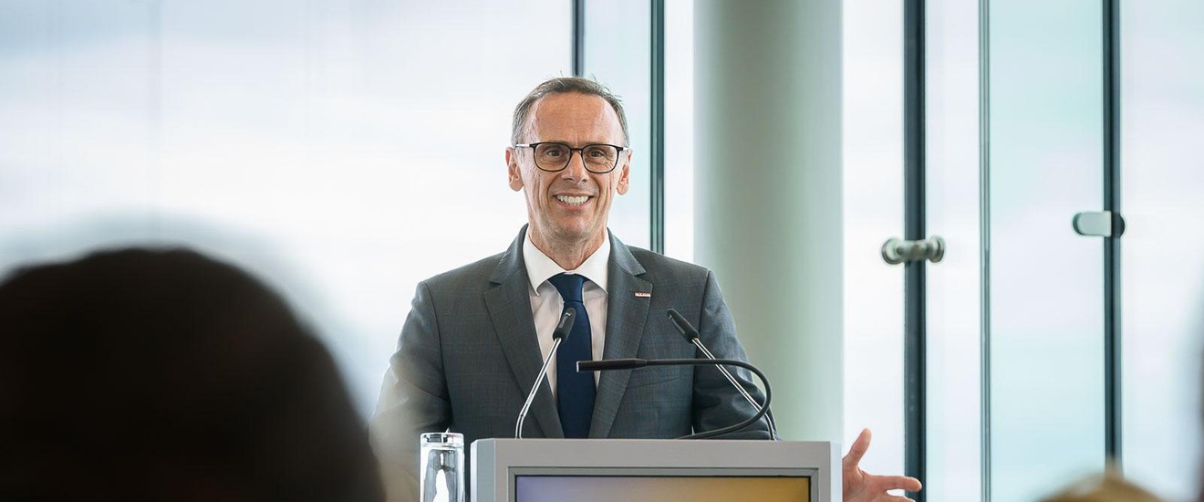 Eine Person mit Sakko, Krawatte, hellem Hemd und Brille steht hinter einem Pult und spricht in eine Mikrofon. Auf dem Pult steht Handelstag. Der Person gegenüber sitzt ein Publikum