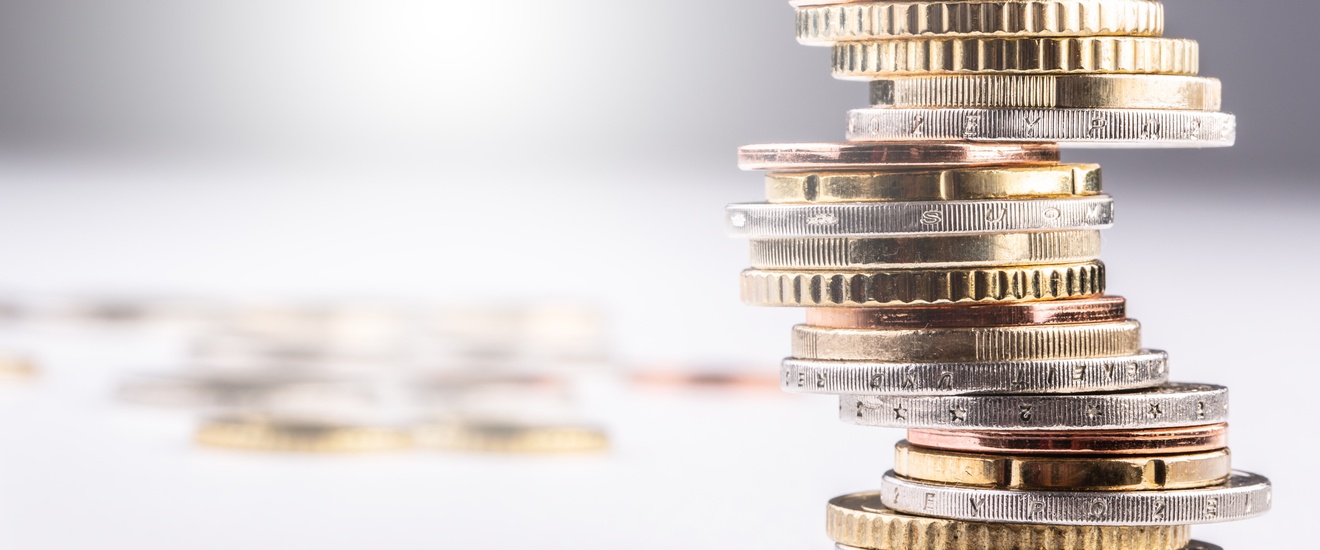 In der rechten Bildhälfte ist ein nicht gleichmäßig gestapelter Münzstapel, der auf einer hellen Oberfläche steht. Im Hintergrund verschwommen liegen weitere Münzen einzeln verstreut.