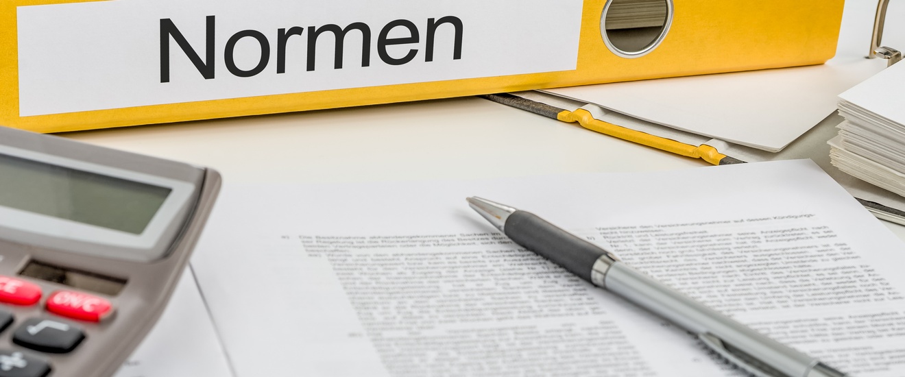 Gelber Ordner mit Beschriftung Normen auf Längsseite liegend, davor Stift auf Dokument und Ausschnitt eines Taschenrechners