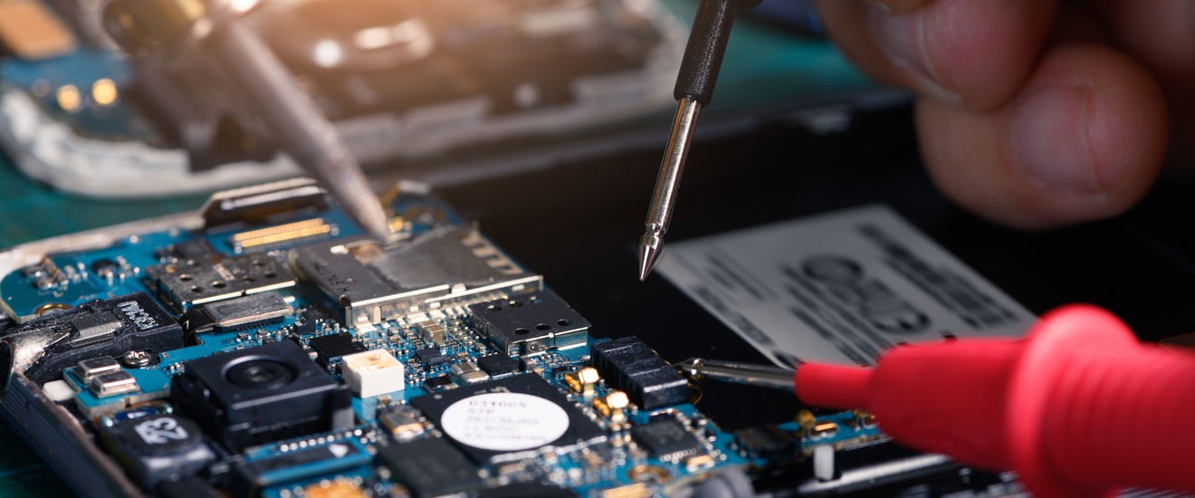 Nahaufnahme einer geöffneten elektronischen Baugruppe. Zwei Messspitzen berühren Bauteile auf einer Leiterplatte. Mehrere elektronische Komponenten, Steckverbindungen und Mikrochips sind auf der Leiterplatte