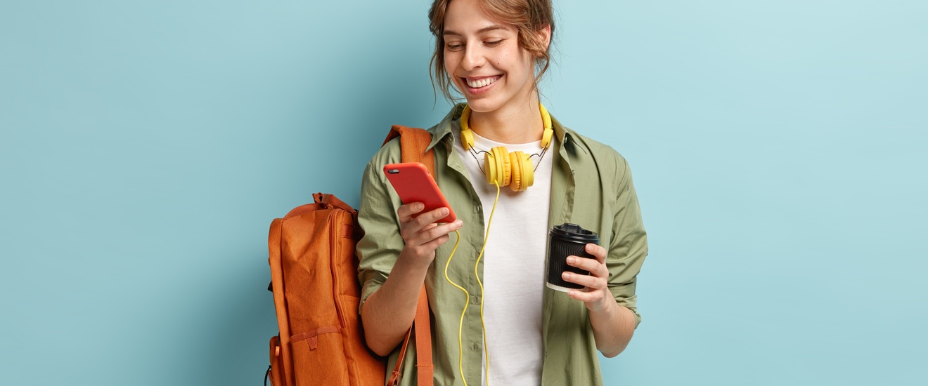 Jugendliche Person mit braunen hochgeschlossenen Haaren, weißem Shirt und grüner offener Bluse blickt auf ihr Smartphone und trägt einen orangen Rucksack sowie gelbe Kopfhörer rund um den Hals und einen Kaffeebecher in der Hand