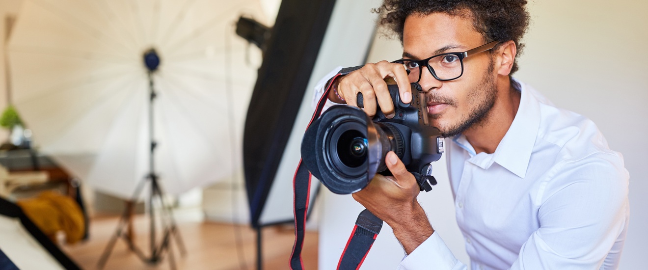 Eine Person mit Brille, in einem weißen Hemd beugt sich mit dem Oberkörper und dem Kopf leicht nach vorne. Sie hält mit beiden Händen seitlich eine Kamera. Die Person blickt geradeaus