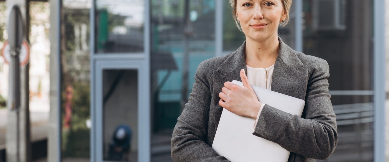 Porträt einer sanft lächelnden Person die zugeklapptes Notebook umfasst hält, im Hintergrund verschwommen Glasfassade eines Gebäudes