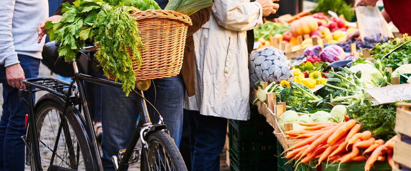 Mehrere Personen stehen um Markstand mit Gemüse, daneben ein Fahrrad mit Gemüse im Korb
