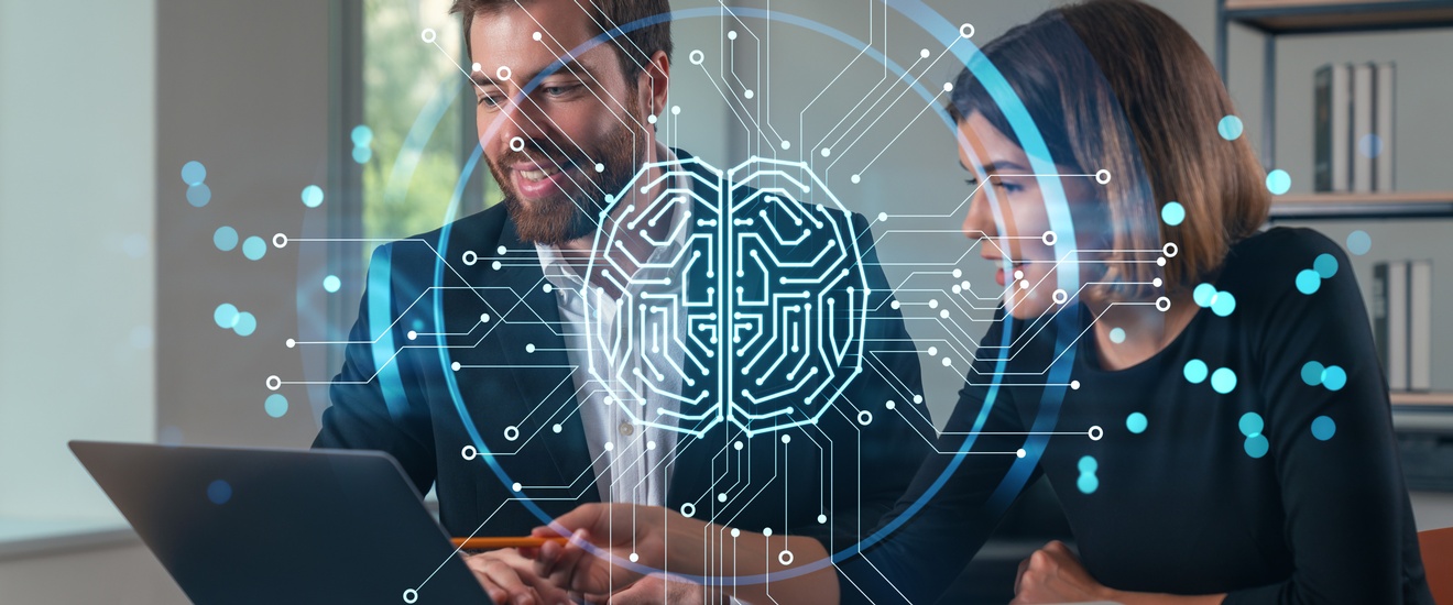 Zwei Personen sitzen hinter einem Tisch. Vor ihnen ist ein aufgeklappter Laptop. Beide blicken darauf. Über dem Bild ist eine Grafik eines Kreises, in dem verschiedene Linien und Punkte sind, die ein Gehirn darstellen.