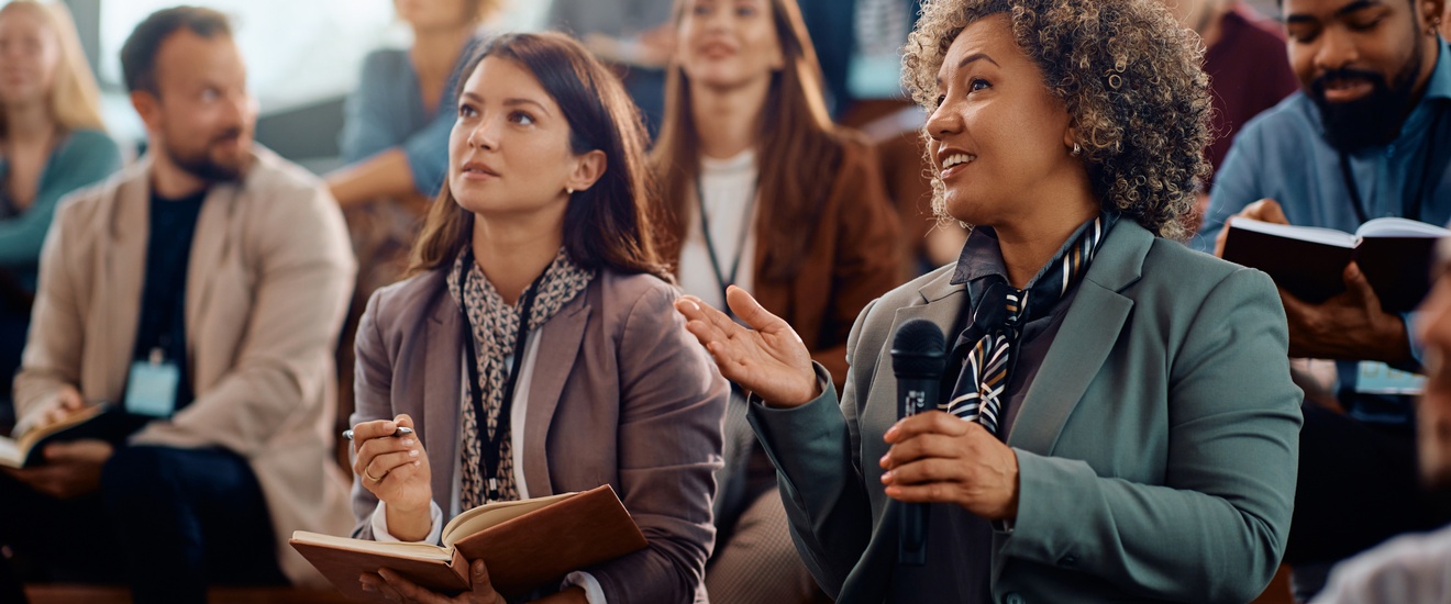 Mehrere Personen in Bürokleidung sitzen in einem Auditorium und blicken nach vorne. Die Person rechts unten im Bild hält in der linken Hand ein Mikrofon