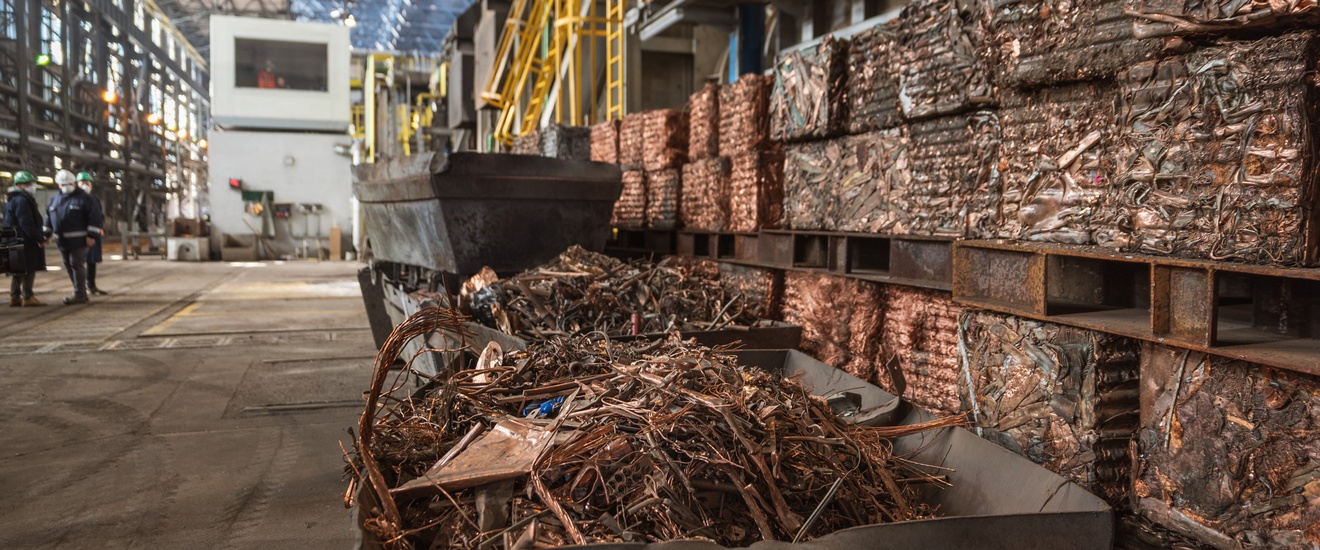 Kupferschrott in einem Container bereit für Recycling in einer Fabrikhalle, im Hintergrund stehen drei Personen in Schutzbekleidungen