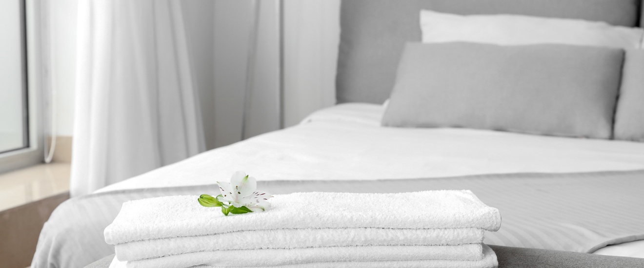 Weiße Handtücher liegen auf einer grauen Kleiderablage vor einem weiß-grau bezogenem Bett