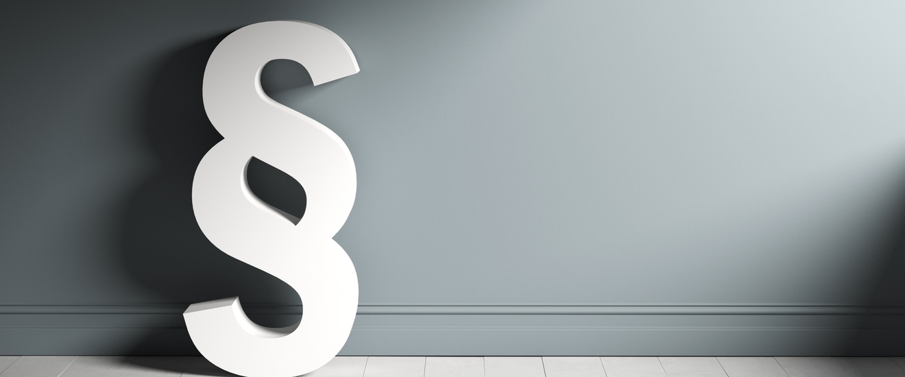 Weißes Paragrafenzeichen lehnt an grauer Wand auf hellem Parkettboden stehend