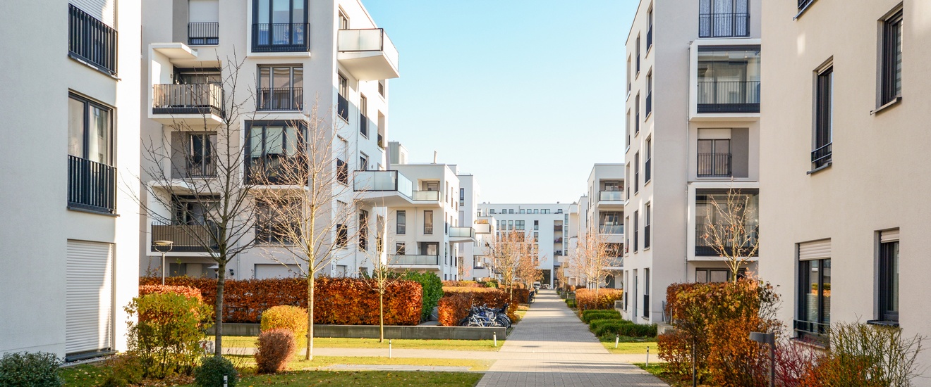 Außenansicht in Reih und Glied angeordneter heller, mehrstöckiger Wohnhäuser mit Balkonen in einer Anlage. Zwischen den Häusern sind asphaltierte Gehwege sowie grüne Streifen mit Bäumen ohne Blättern und Büschen