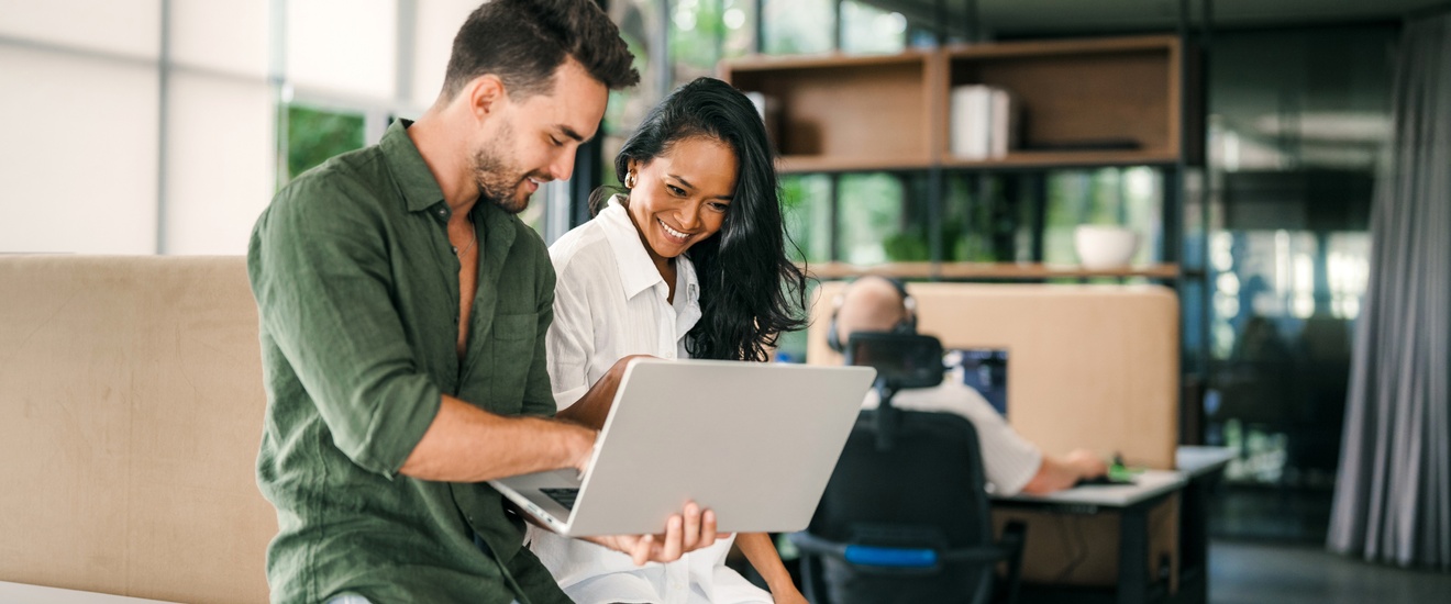 Zwei Personen sitzen nebeneinander auf einem Tisch in einem Büro. Die linke Person hält einen aufgeklappten Laptop. Die rechte Person blickt auf den Bildschirm des Laptops. Im Hintergrund sitzt eine Person an einem Schreibtisch