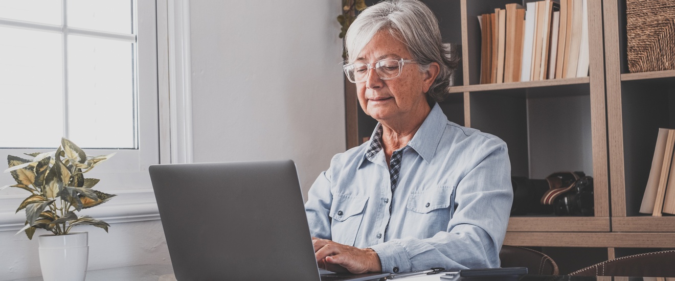 Person mit grauen Haaren und Brille sitzt an Tisch und blickt auf Laptop