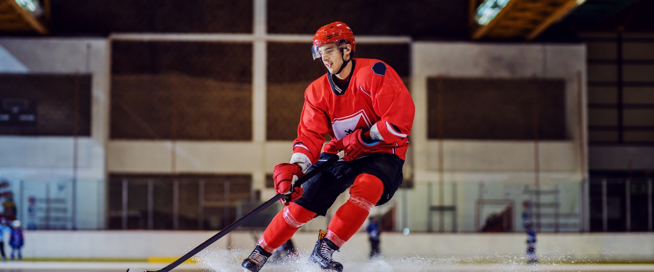 Eine Person in Eishockey-Ausrüstung fährt mit Schlittschuhen auf einer Eisfläche in einer Halle und manövriert einen Puck mit einem Schläger.
