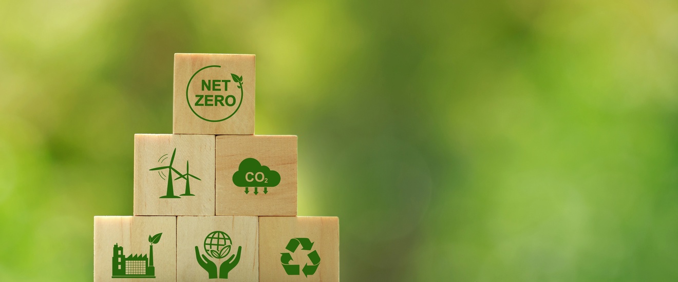 Mehrere Holzwürfel aufeinander gestapelt auf Erde mit grünen Symbolen zum Thema Klimaschutz, oberster Würfel mit Schriftzug Net Zero, Hintergrund verschwommen grün 