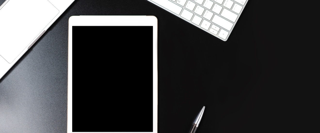 Mock-up von einem Tablett auf einem schwarzen Untergrund neben einem Laptop, einer Tastatur sowie einem Kugelschreiber, Topshot