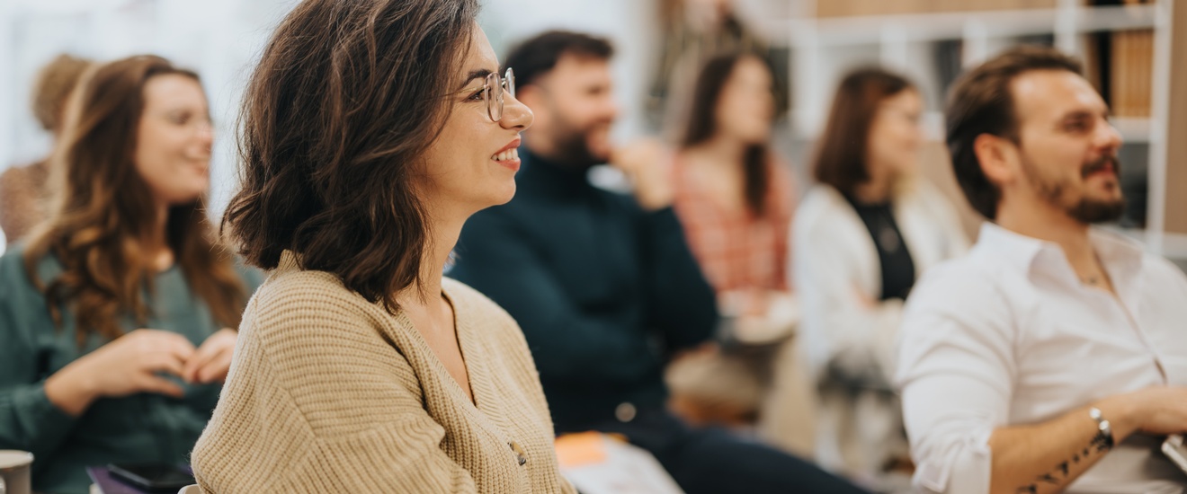 In mehreren Sesselreihen sitzen hintereinander mehrere Personen. Alle Personen blicken leicht nach rechts. Im Hintergrund sind eine Fensterfront und ein Regal mit Büchern