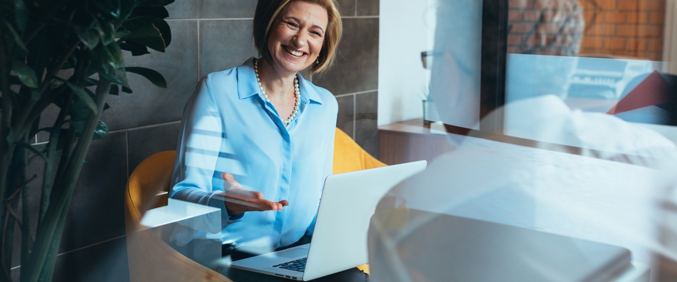 Lächelnde Person sitzt in gelbem Stuhl mit aufgeklappten Notebook am Schoß und unterhält sich mit anderer Person in Rückenansicht