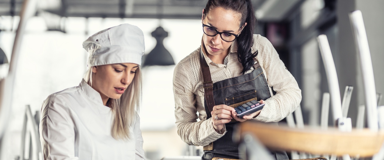 Frontalaufnahme einer an einem Tisch sitzenden Person in weißer Kleidung und mit weißer Mütze auf dem Koch. In den Händen hält sie ein Tablet, auf das sie blickt. Rechts daneben steht eine weitere Person mit Schürze, die auch auf das Tablet blickt