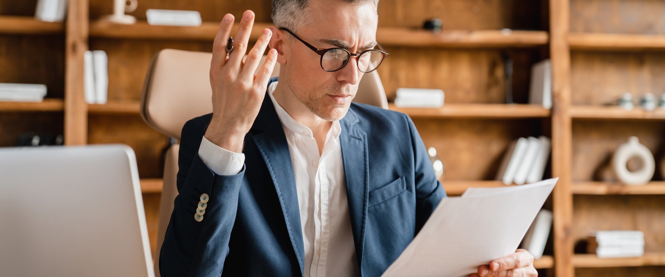 Person im Anzug trägt eine Brille und sitzt mit fragendem Blick an einem Schreibtisch mit Computer und hält Papier in einer Hand ein Papier und in der anderen einen Stift, im Hintergrund steht ein braunes Holzregal mit Büchern und Dekoelementen