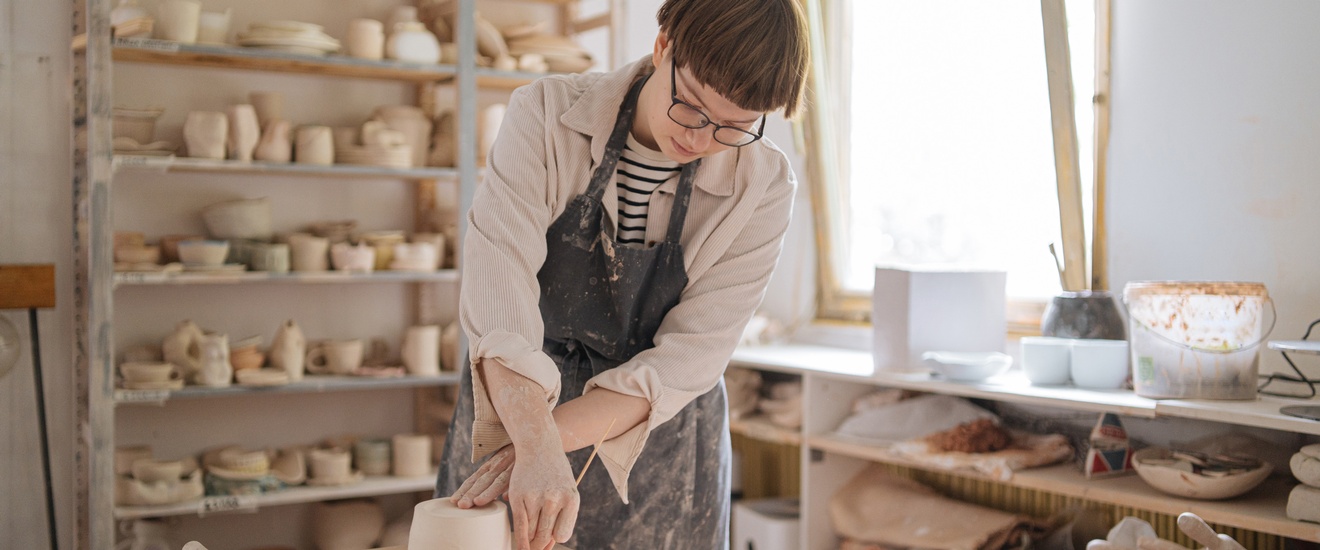 Person mit Brillen in Schürze steht an Tisch in Töpfereiwerkstatt und bearbeitet Ton mit Holzstäbchen, im Hintergrund Regale mit Töpferwaren