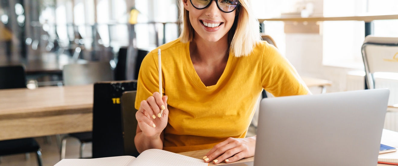 Lächelnde Person mit Brille und erhobenem Stift in der Hand sitzt vor aufgeklapptem Laptop und Büchern 