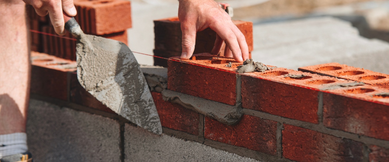 Eine Person legt einen Ziegel auf eine bereits ein paar Zentimeter hohe Mauer. In der rechten Hand hält die Person einen Spachtel