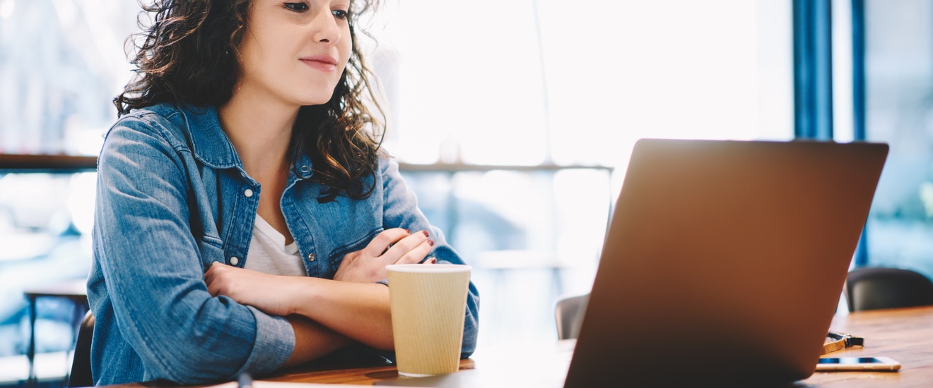 Schmunzelnde Person mit verschränkten Armen sitzt vor Laptop, daneben Block mit Stift und Pappbecher