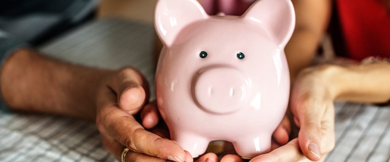 Mehrere Hände halten ein Rosa Sparschwein, darunter liegen Geldmünzen auf einem Tisch