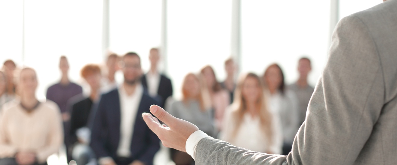 Die Rückenansicht einer Person im Anzug ist im rechten Bildrand. Die Person hat den linken Arm von sich weggestreckt. Gegenüber der Person sitzen viele Leute auf Stühlen in einem Raum