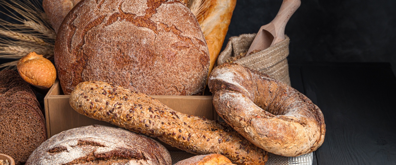 Auf einem dunklen Holzuntergrund liegen verschiedene Backwaren in einer Kiste und davor, zum Beispiel Brot, Baguette oder Weckerl. In der Kiste sind außerdem die oberen Teile einer Getreidepflanze. Vor den Backwaren verstreut liegen Körner.