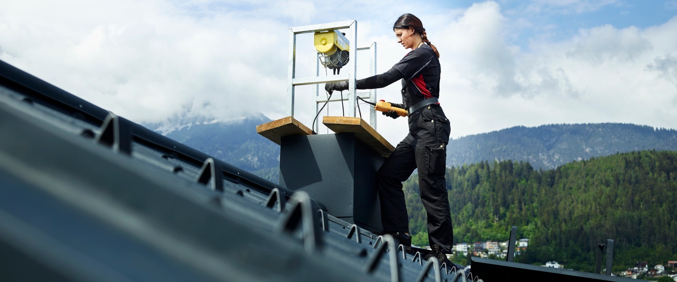 Eine Frau arbeitet an einem Kamin. Im Vordergrund das schwarze Schrägdach. Im Hintergrund eine Bergkulisse