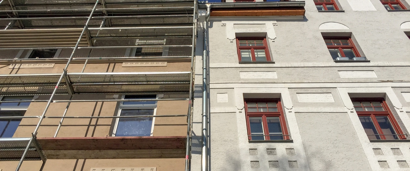 Die Fassaden zweier Altbaugebäude direkt nebeneinander. Vor der Fassade des linken Hauses ist ein Baugerüst. Über den Gebäuden ist der blaue Himmel