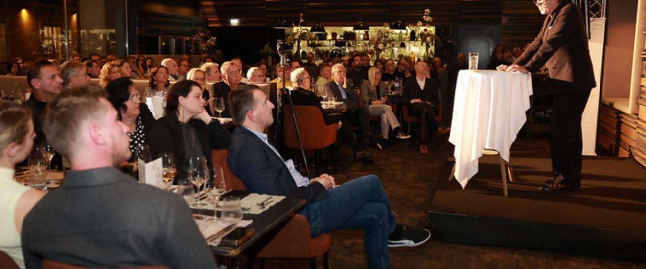 Eine größere Gruppe von Menschen hört einem Vortragenden zu, der auf einer kleinen Bühne an einem Stehtisch spricht.