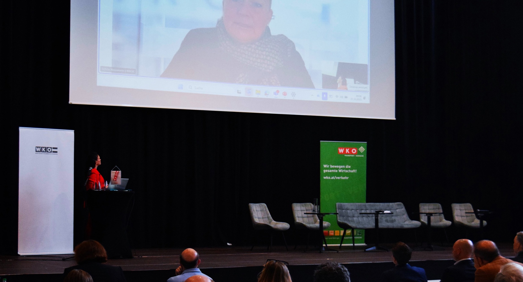 Rückenansicht mehrerer Personen, die in einem Raum an Tischen sitzen und alle zu einer Leinwand blicken, die an der Wand montiert ist und auf der eine Person zu den Personen spricht