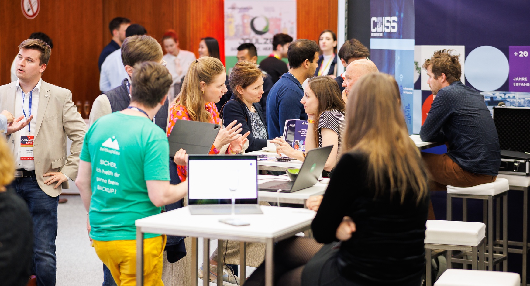 Mehrere Menschen sitzen im Rahmen einer Veranstaltung auf Hochstühlen an kleinen Tischen nebeneinander, auf den Tischen stehen aufgeklappte Laptops