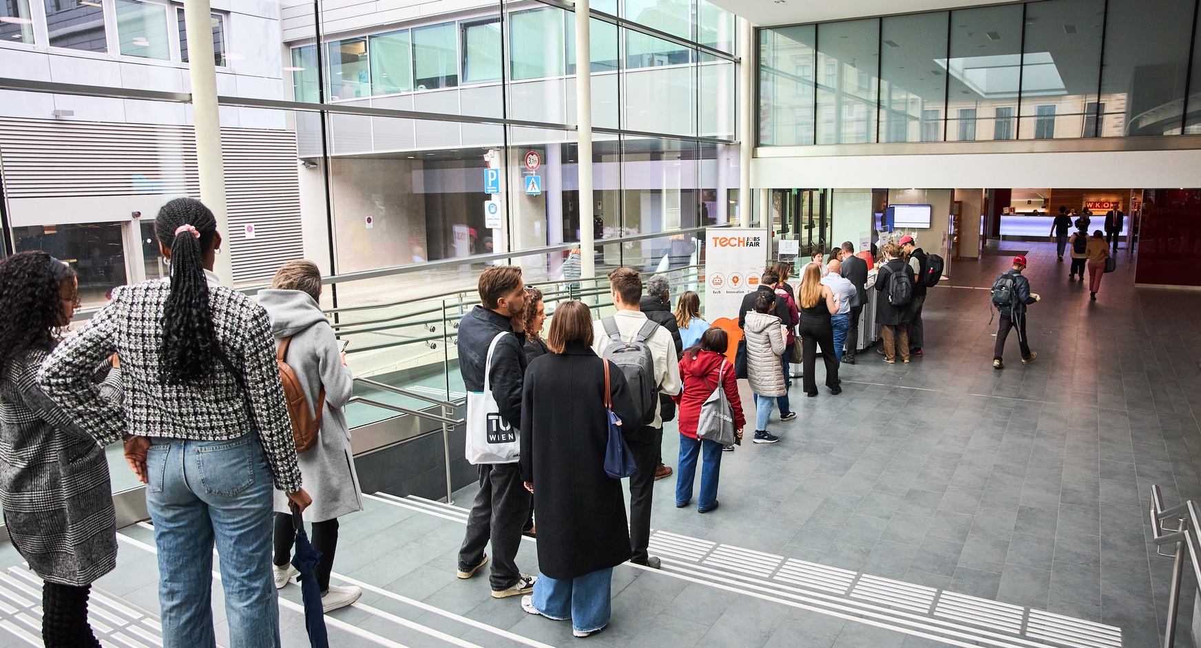 Menschen stehen in einer Reihe nebeneinander und sind von hinten fotografiert. Sie warten im Foyer auf den Check-In einer Veranstaltung 