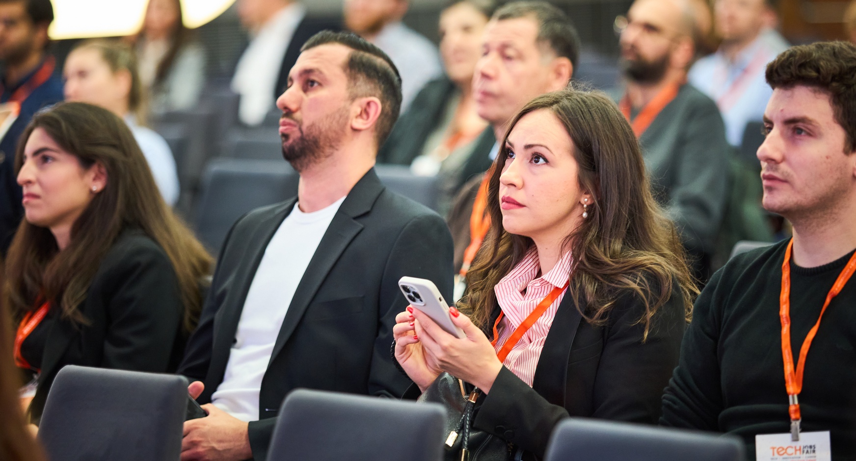 Nahaufnahme von Personen, die nebeneinander auf Sesseln sitzen. Eine Person hält ein Smartphone in der Hand