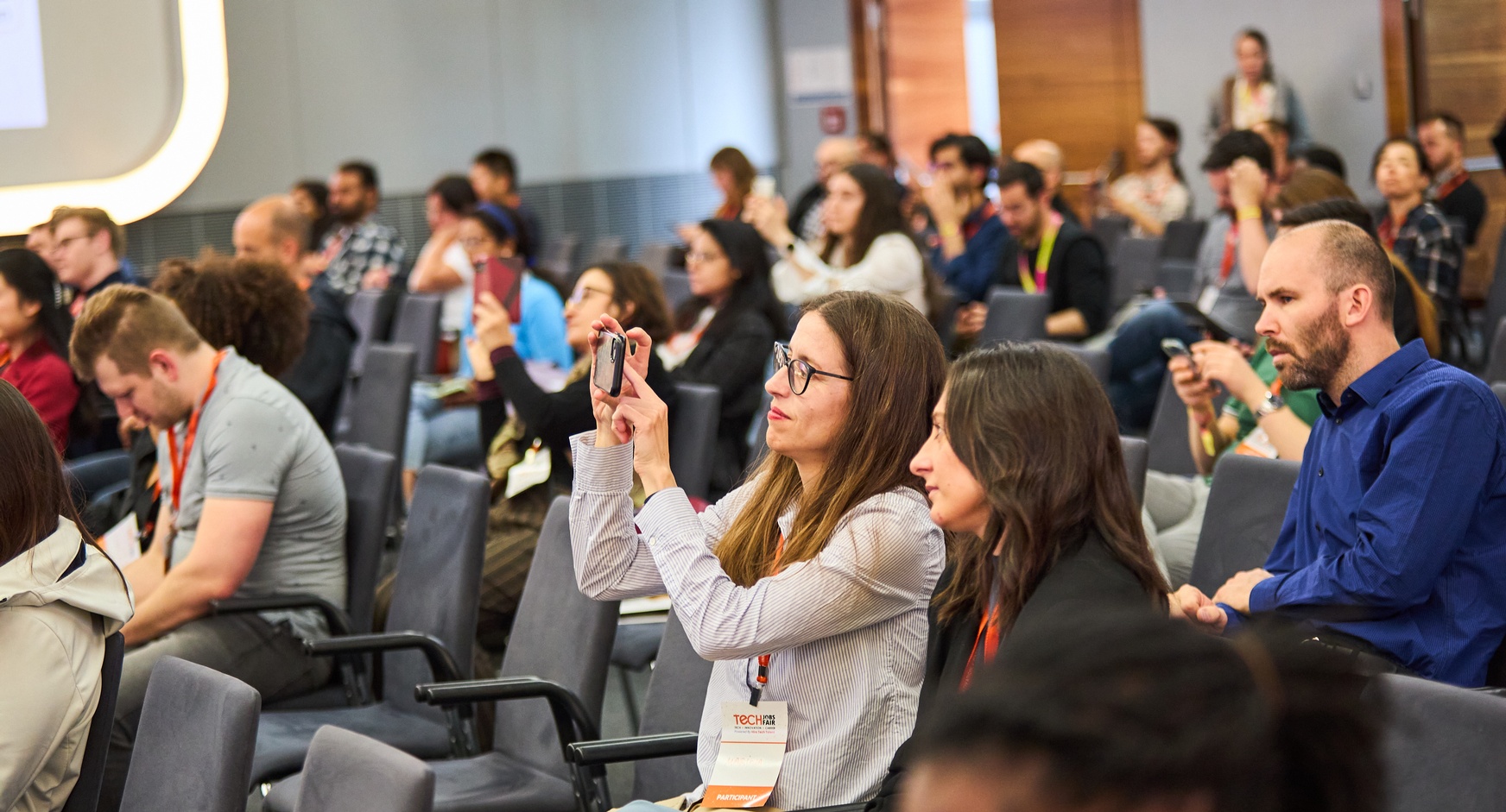 Personen sitzen nebeneinander auf Sesseln, die in Reihen aufgestellt sind. Eine Person macht ein Selfie mit der Kamera ihres Smartphones 