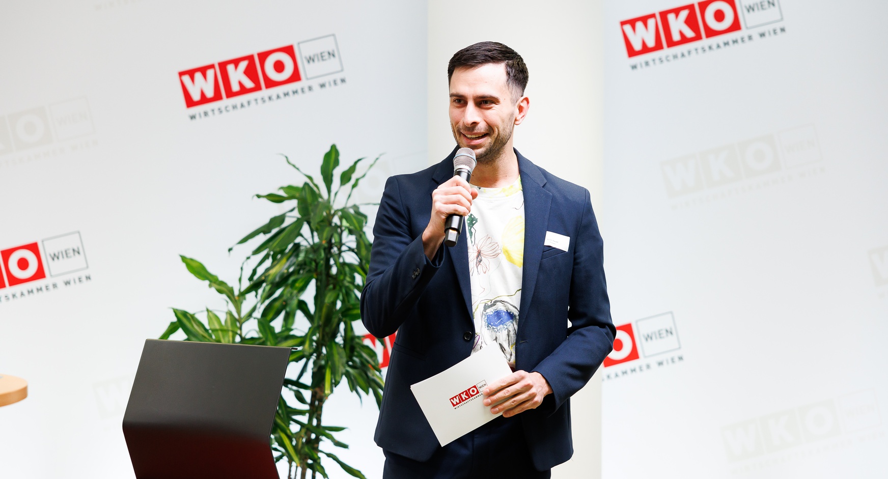 Stehende Person spricht in Mikrofon vor großer Wand mit rotweißem Logo WKO und Schriftzug Wirtschaftskammer Wien, hält in einer Hand Kärtchen mit rotweißem Logo WKO, daneben rotschwarzer Sessel und Zimmerpflanze