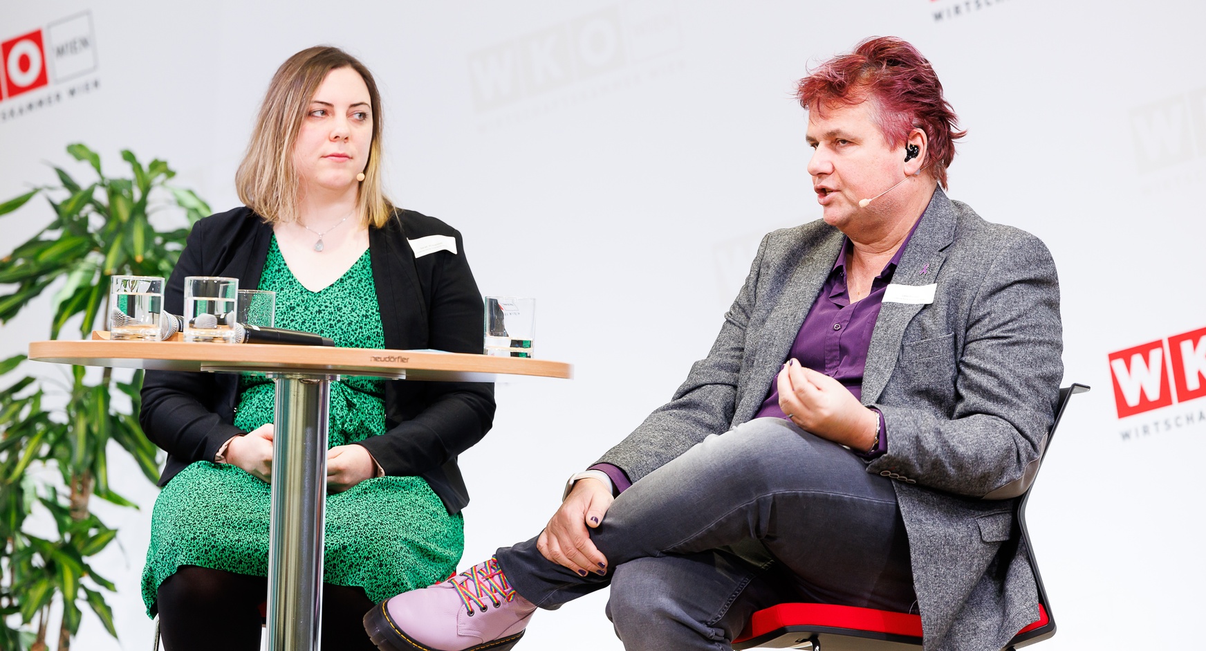 Zwei Personen mit Headmikrofonen sitzen auf erhöhten Stühlen neben Stehtisch auf Bühne vor Wand mit rotweißen Logos WKO und Schriftzügen Wirtschaftskammer Wien, eine der Personen spricht