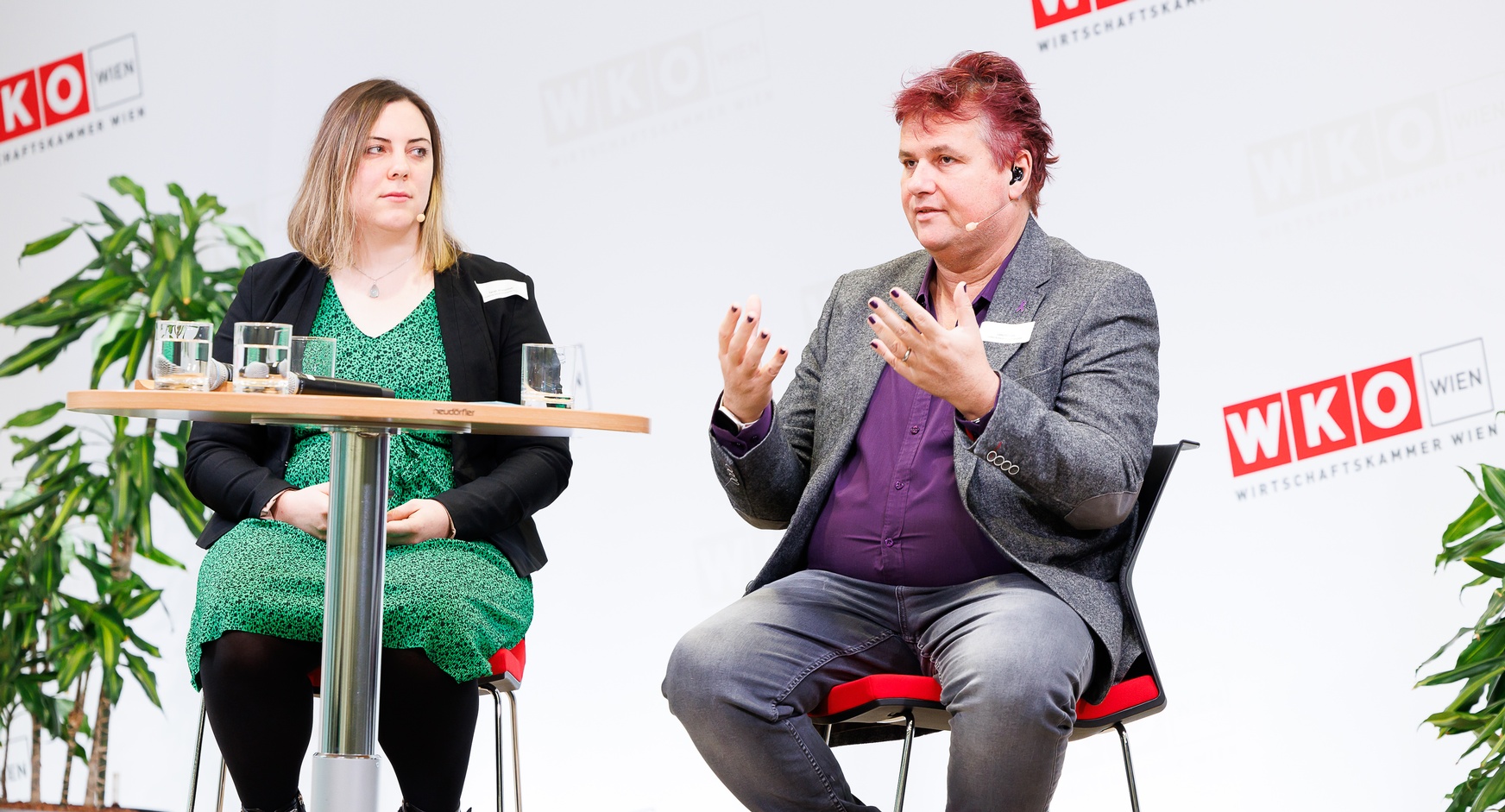 Zwei Personen mit Headmikrofonen sitzen auf erhöhten Stühlen neben Stehtisch auf Bühne vor Wand mit rotweißen Logos WKO und Schriftzügen Wirtschaftskammer Wien, eine der Personen spricht und hält Hände vor sich in die Luft