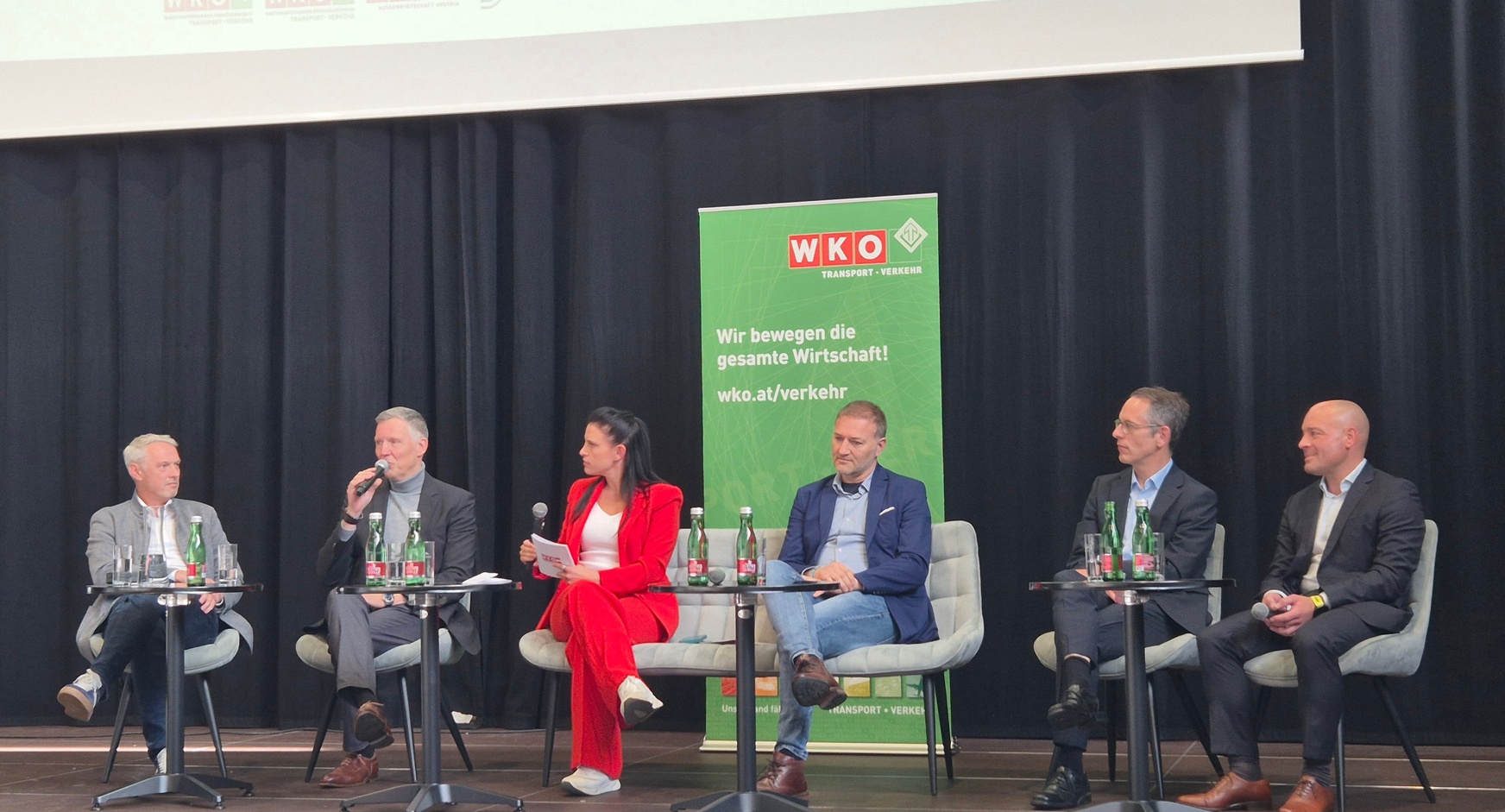 Auf einer Bühne sitzen nebeneinander auf sechs Stühlen sechs Personen. Eine der Personen hält ein Mikrofon und spricht in dieses. Hinter ihnen ist ein Banner, auf dem Wir bewegen die gesamte Wirtschaft steht