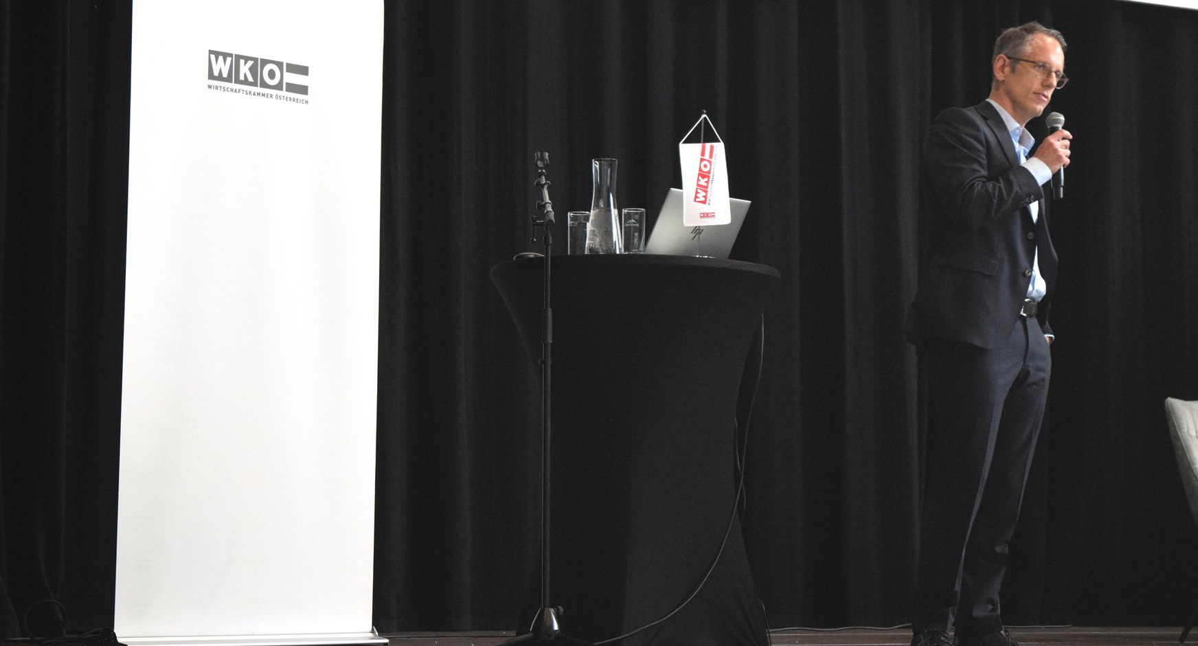 Eine Person steht neben einem Rundtisch, auf dem ein Laptop steht und ein Mikrofon liegt. Die Person spricht in ein Mikrofon. Hinter der Person ist ein weißer Banner, auf dem Wirtschaftskammer Österreich steht