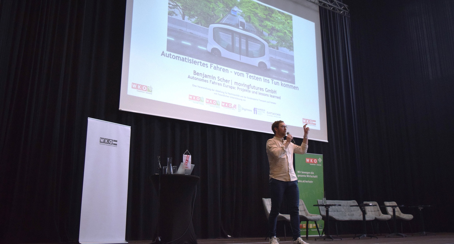 Eine Person steht neben einem Rundtisch, auf dem ein Laptop steht und ein Mikrofon liegt. Die Person spricht in ein Mikrofon. Hinter der Person ist ein weißer Banner, auf dem Wirtschaftskammer Österreich steht. Hinter der Person ist eine Leinwand, auf der automatisiertes Fahren - vom Testen ins Tun kommen steht