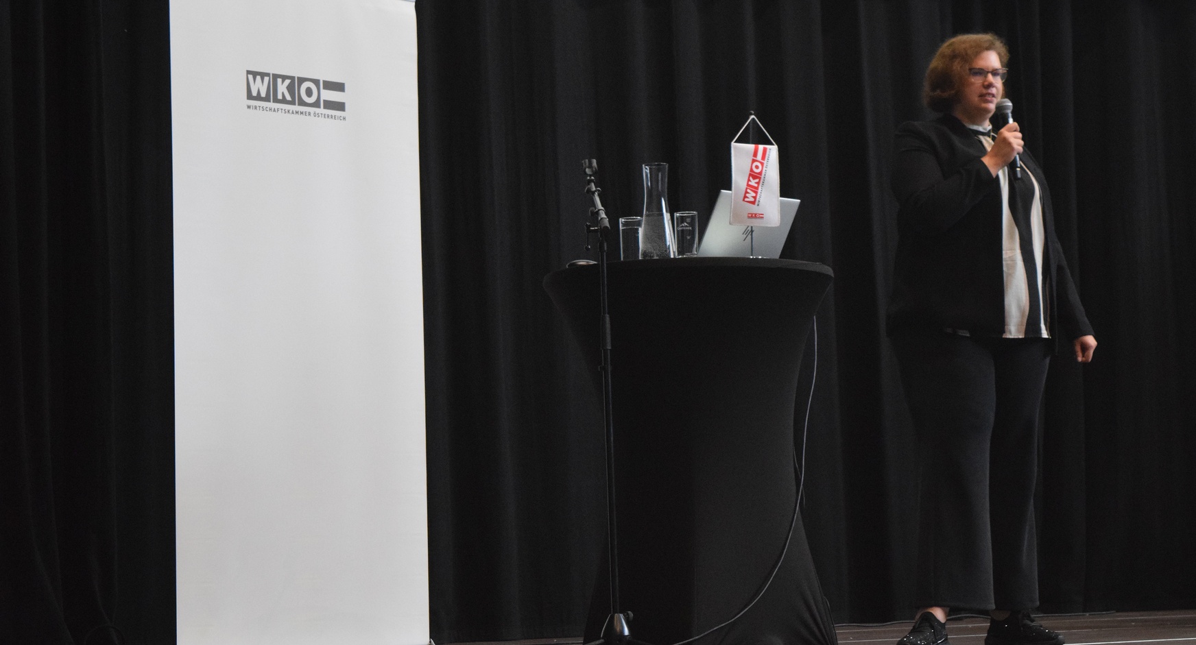 Eine Person steht neben einem Rundtisch, auf dem ein Laptop steht und ein Mikrofon liegt. Die Person spricht in ein Mikrofon. Hinter der Person ist ein weißer Banner, auf dem Wirtschaftskammer Österreich steht